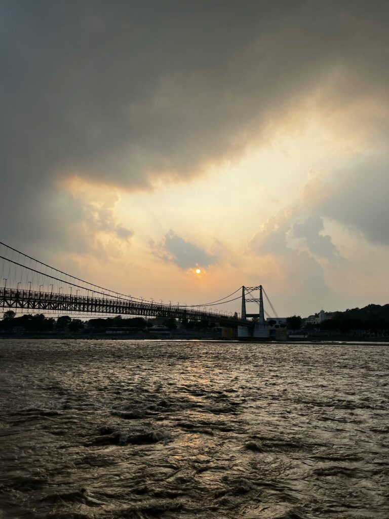 Captured during sunset, this image showcases a suspension bridge over a flowing river with dramatic clouds.