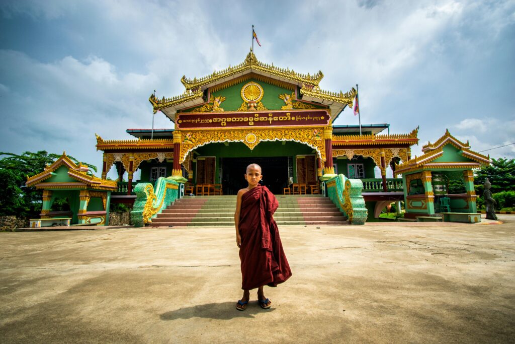 A young monk stands before a traditional Burmese temple on a sunny day.